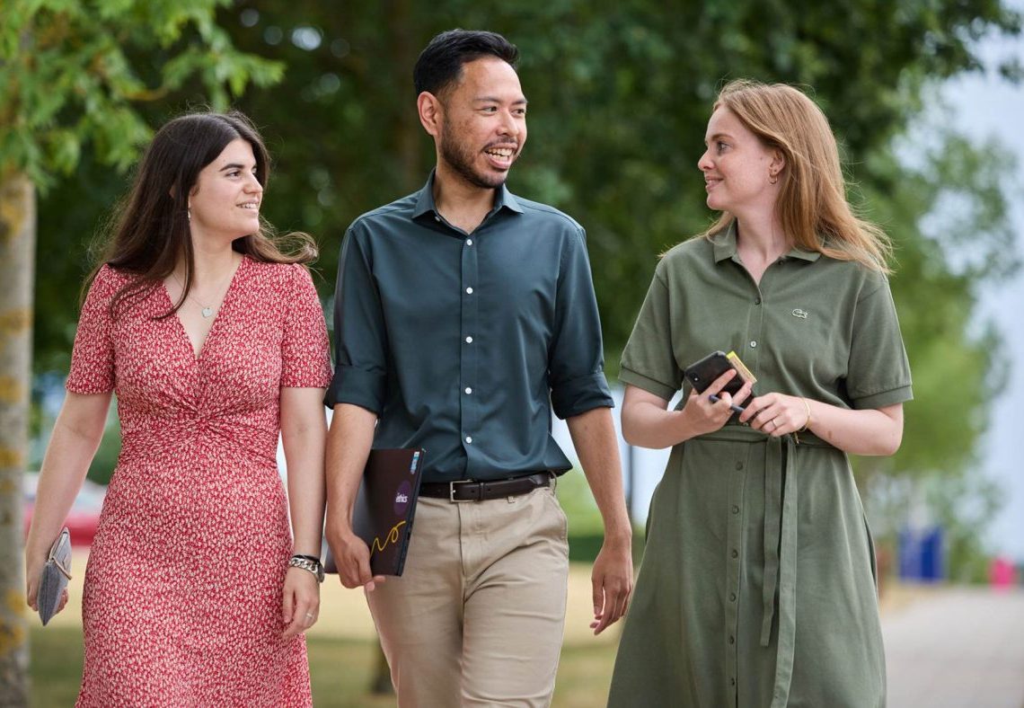 Three colleagues walking and talking outdoors under green trees.
